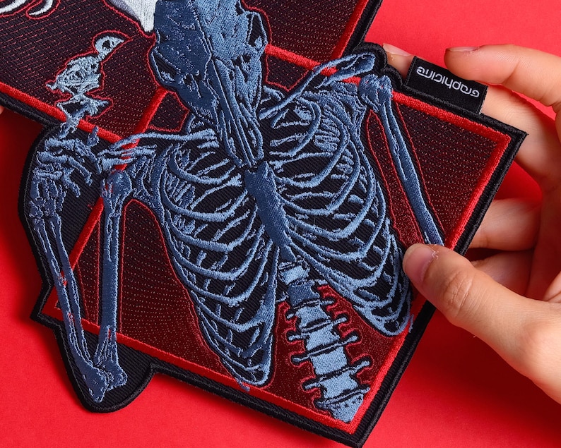 May include: Embroidered patch featuring a blue skeletal design on a black background, framed by a red border. The patch has a diamond shape with a small tag that reads "graphicine". The design includes a detailed depiction of a rib cage and spine.