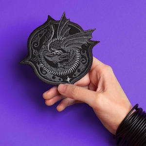 May include: A black embroidered patch featuring a dragon design within a decorative shield shape. The dragon is depicted with detailed wings and scales. The patch is held against a purple background.