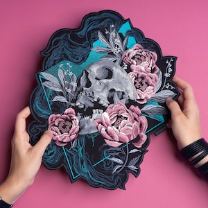 May include: Embroidered patch featuring a gray skull surrounded by pink peonies and foliage on a black background. The patch has a unique, irregular shape with teal accents and is held against a pink backdrop.