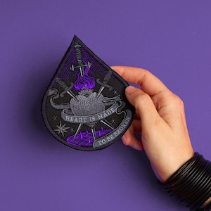 May include: A black and purple embroidered patch with a heart pierced by swords. The patch reads "HEART IS MADE TO BE BROKEN" in gray text. The design includes purple flames and star details against a black background.