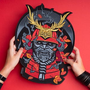 May include: A large embroidered patch featuring a stylized samurai mask in shades of grey and red, topped with a golden owl-like helmet crest. The design includes swirling grey smoke, scattered orange leaves, and a black circular background border.