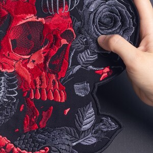 以下が含まれることがあります： 鮮やかな赤い頭蓋骨が中心に刺繍された大きなワッペンで、ダークグレーの蛇が絡み合い、詳細なダークグレーのバラと葉で囲まれています。すべて無地の黒い布地を背景にしています。