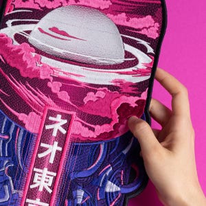 Peut inclure: Écusson brodé représentant une planète avec des anneaux, sur fond de nuages roses et d'un motif violet et bleu. L'écusson comprend du texte japonais dans un rectangle rouge vertical. L'écusson est tenu sur un fond rose vif.