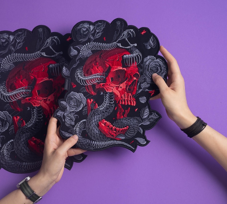 以下が含まれることがあります： 黒い刺繍ワッペン。赤い頭蓋骨に灰色の骸骨の蛇が絡みつき、周囲を灰色のバラが囲むデザイン。黒い背景にスカラップ状の縁取りが施され、精巧な刺繍糸の質感が特徴的です。