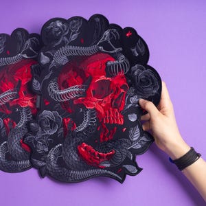 以下が含まれることがあります： 鮮やかな赤い頭蓋骨と詳細な灰色のヘビの骨格が絡み合った、黒い刺繍入りのダイカットワッペン2枚。黒い生地の上に、中央の頭蓋骨とヘビのモチーフを囲むように、濃い灰色のバラと葉がデザインされています。