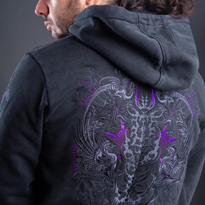 May include: A charcoal gray hooded sweatshirt featuring an intricate, symmetrical embroidery design on the back. The embroidery showcases stylized avian figures and ornate patterns in shades of dark gray and vibrant purple thread.