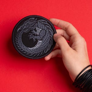 May include: A round, black patch featuring a gray dragon in a circular design. The dragon is depicted with intricate details, including scales and claws. The patch is held against a red background.
