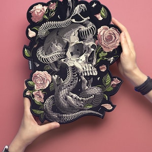 May include: Black patch featuring a skull intertwined with a snake, surrounded by pink roses and green leaves. The patch has a unique, irregular shape and is held against a pink background.