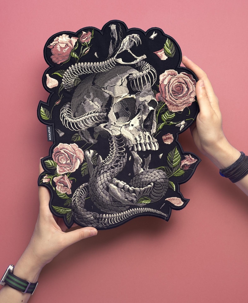 May include: Black patch featuring a skull intertwined with a snake, surrounded by pink roses and green leaves. The patch has a unique, irregular shape and is held against a pink background.