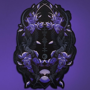 May include: A black embroidered patch with a symmetrical design featuring two crows with outstretched wings, purple roses, and a central vertical element. The patch has a scalloped edge and is set against a purple background.