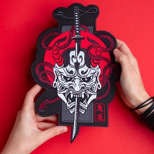 May include: Embroidered patch featuring a white Oni mask with red horns and a katana sword. The design includes a red sun, red rope-like accents, and Japanese text. The patch is held against a red background.