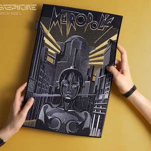 May include: A black embroidered patch with gold stitching depicting a futuristic cityscape with the word "Metropolis" in a stylised font. The patch features a woman's face in the centre.