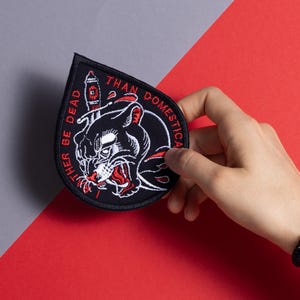 Può includere: Toppa ricamata in bianco e nero con una testa di pantera e un pugnale. Il testo intorno alla pantera dice "Rather be dead than domesticated".