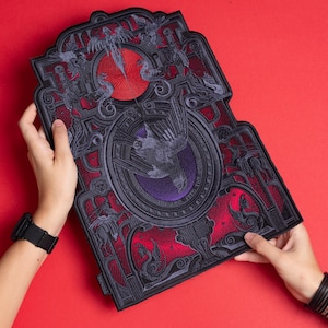 May include: A decorative, rectangular patch with a gothic design. The patch features a central bird motif, surrounded by ornate, dark grey and red filigree. A red circle and purple accents add colour to the design.
