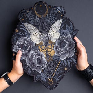 Può includere: Toppa ricamata con un design dettagliato di una cicala con le ali spiegate, circondata da peonie grigie e fogliame. La toppa ha uno sfondo nero con accenti dorati e grigi e un bordo decorativo. La toppa è tenuta da due mani.