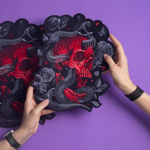 May include: Two black decorative patches with a red skull, gray snakes, and roses design. The patches have a scalloped edge and are held against a purple background.