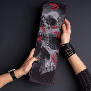 May include: A rectangular black fabric patch featuring detailed grey and white embroidery of two stacked skulls. The design is accented with small, vibrant red centipedes crawling across the skulls. The entire piece is bordered by a thin red stitch.