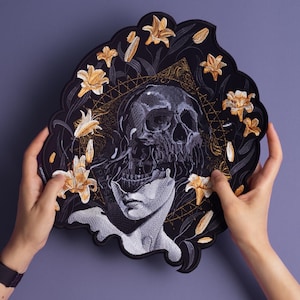 May include: Embroidered patch featuring a skull with a floral design. The skull is surrounded by white lilies and a black background. The patch is made of fabric and has a detailed design.