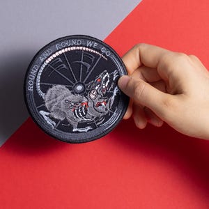May include: Round, black embroidered patch with a rat and wheel design. The patch has the words "ROUND AND ROUND WE GO" in white lettering around the edge. The rat is gray and the wheel is black.