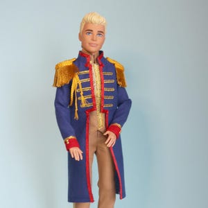 May include: A doll dressed in a blue coat with gold epaulettes and trim, red details, and gold buttons. The doll also wears tan trousers and brown shoes. The doll has blonde hair and stands against a light blue backdrop.