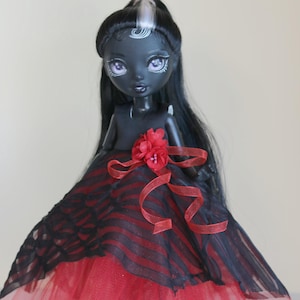 May include: A custom doll with dark skin, black hair, and a red and black dress. The doll has a red flower accent and a red ribbon. The dress has a red tulle skirt and a black and red striped bodice.