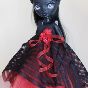 May include: A custom doll with dark skin, black hair, and a striking red and black dress. The doll's dress features a red tulle skirt, a black and red striped bodice, and a red flower accent. The doll has detailed facial features.
