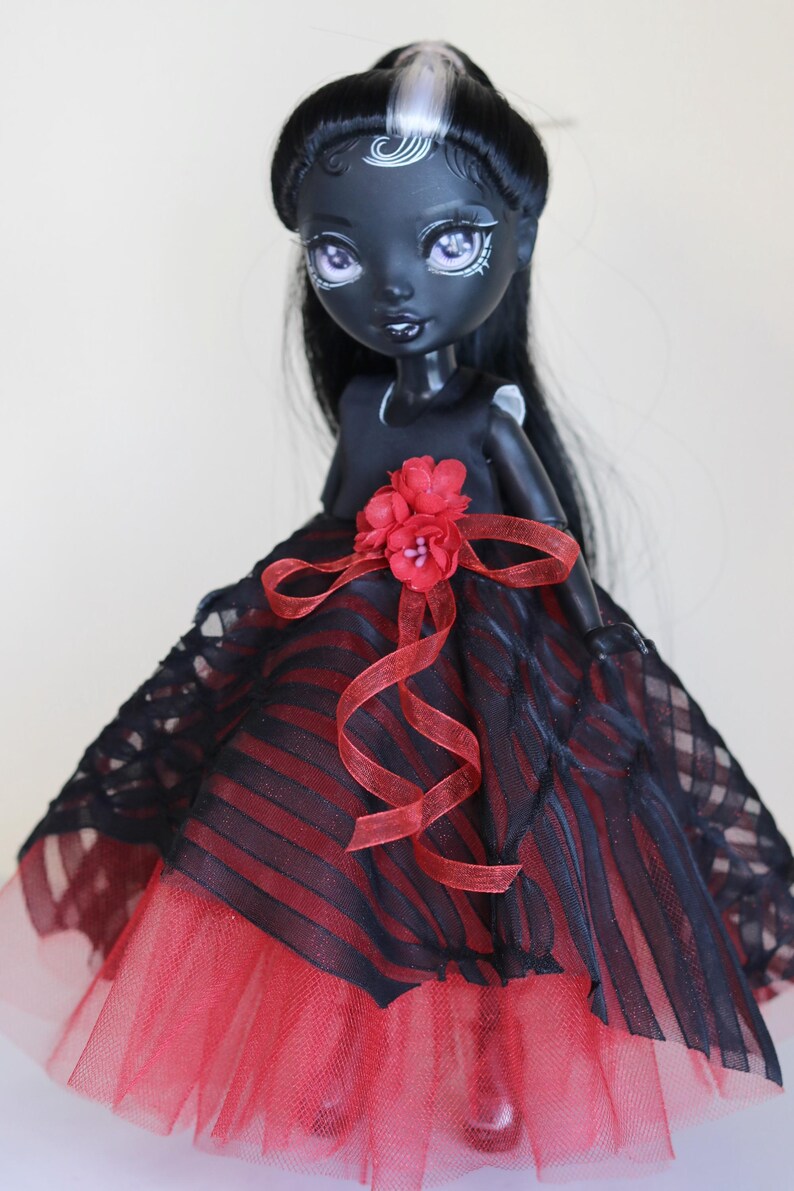 May include: A custom doll with dark skin, black hair, and a striking red and black dress. The doll's dress features a red tulle skirt, a black and red striped bodice, and a red flower accent. The doll has detailed facial features.