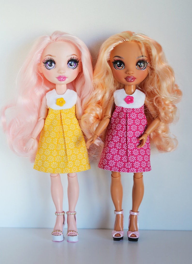 May include: Two dolls wearing floral print dresses. The doll on the left has pink hair and is wearing a yellow dress with white flowers. The doll on the right has brown hair and is wearing a pink dress with white flowers.