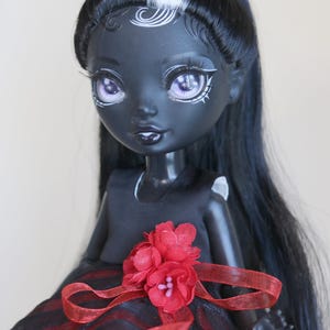 May include: A dark-skinned doll with long black hair, a white streak, and purple eyes. The doll wears a black dress with a red and black striped skirt, accented with a red flower and ribbon.