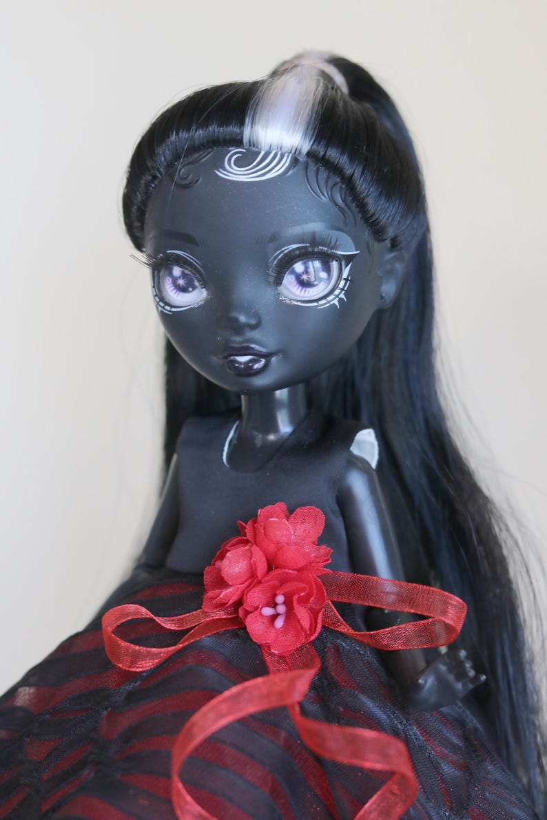 May include: A dark-skinned doll with long black hair, a white streak, and purple eyes. The doll wears a black dress with a red and black striped skirt, accented with a red flower and ribbon.