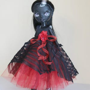 May include: A custom doll with dark skin, black hair, and a striking red and black dress. The doll's dress features a red tulle skirt, a black and red striped bodice, and a red flower accent. The doll has pale purple eyes and is wearing black shoes.