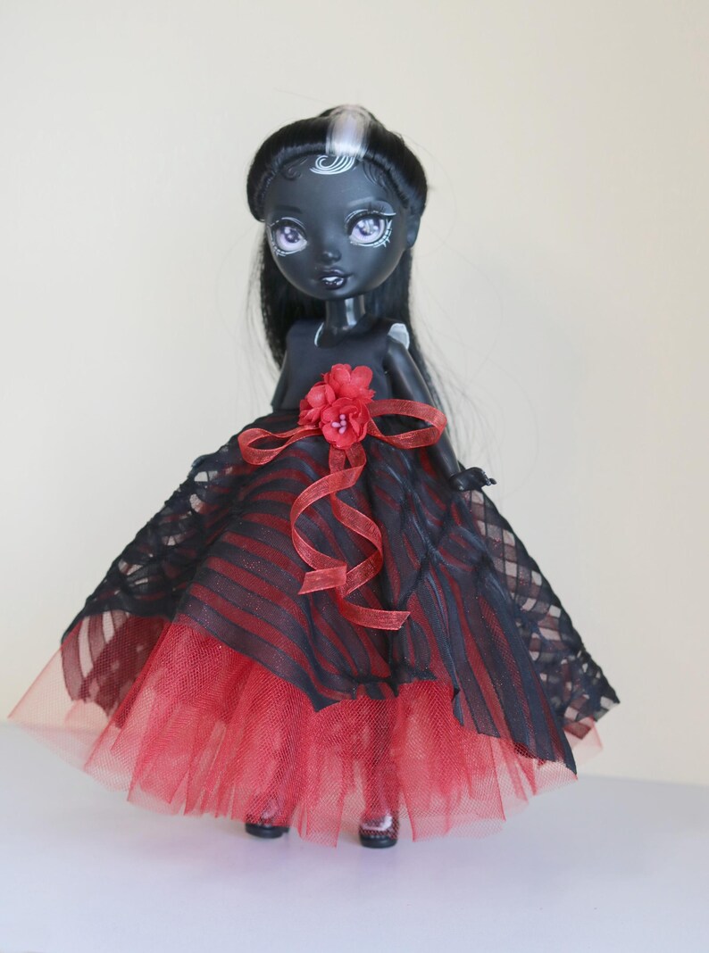 May include: A custom doll with dark skin, black hair, and a striking red and black dress. The doll's dress features a red tulle skirt, a black and red striped bodice, and a red flower accent. The doll has pale purple eyes and is wearing black shoes.