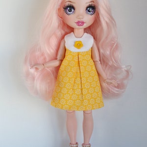 May include: A doll with pink hair wearing a yellow dress with white flowers and a white collar with a yellow flower button. The doll is wearing white platform shoes with pink accents and has purple eyes.