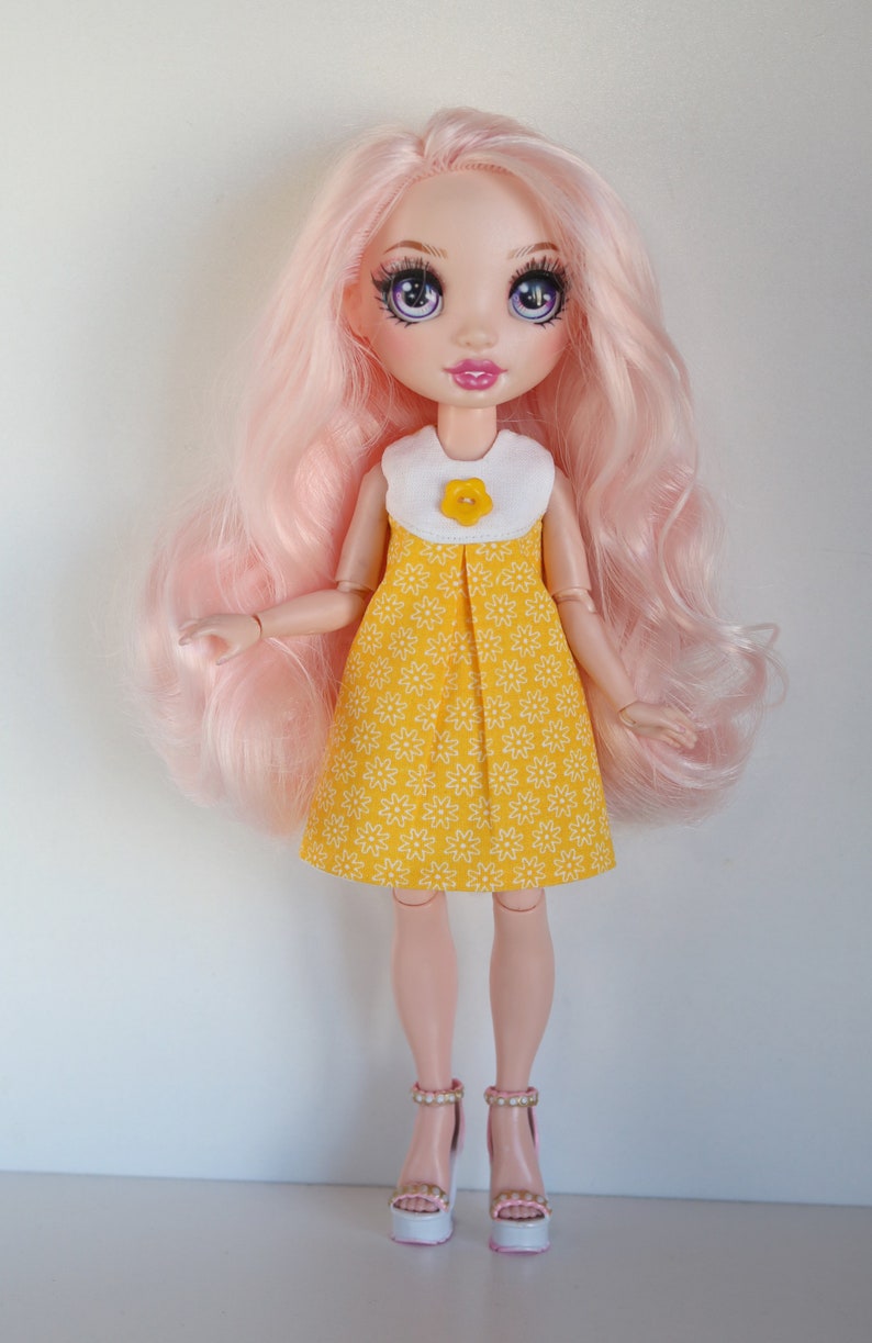 May include: A doll with pink hair wearing a yellow dress with white flowers and a white collar with a yellow flower button. The doll is wearing white platform shoes with pink accents and has purple eyes.