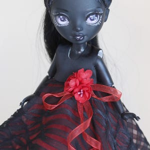 May include: A fashion doll with dark skin, black hair, and striking makeup. The doll wears a black dress with a red and black striped skirt, accented with a red flower and ribbon. The doll's hair has a white streak.