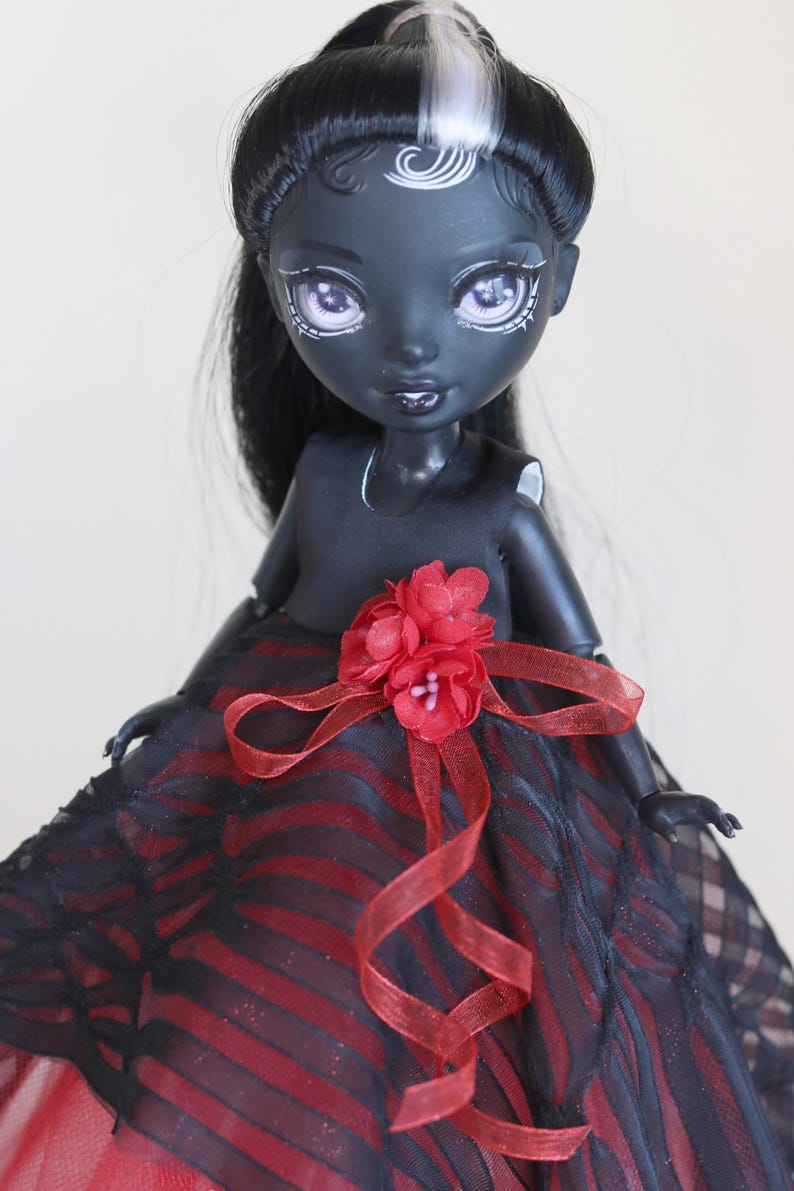 May include: A fashion doll with dark skin, black hair, and striking makeup. The doll wears a black dress with a red and black striped skirt, accented with a red flower and ribbon. The doll's hair has a white streak.