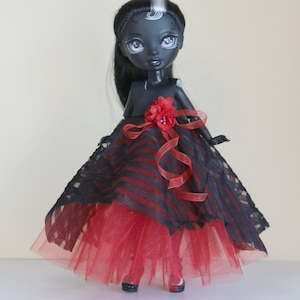 May include: A black doll with long black hair and a white streak, wearing a red and black striped dress with a red tulle skirt. The dress is accented with a red flower and ribbon.