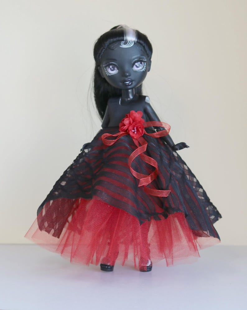 May include: A black doll with long black hair and a white streak, wearing a red and black striped dress with a red tulle skirt. The dress is accented with a red flower and ribbon.