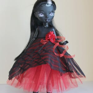 May include: A dark-skinned doll with long black hair and a red and black striped dress. The doll has a red flower on the bodice and red ribbon accents. The skirt is layered with red tulle.
