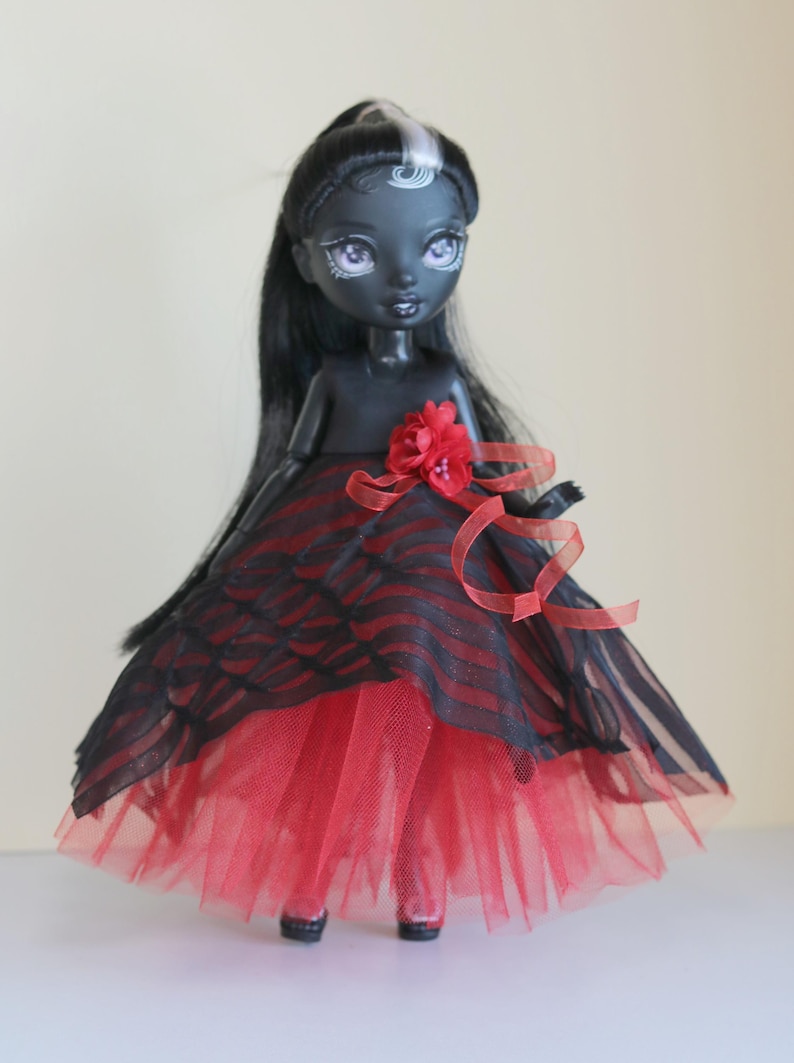 May include: A dark-skinned doll with long black hair and a red and black striped dress. The doll has a red flower on the bodice and red ribbon accents. The skirt is layered with red tulle.