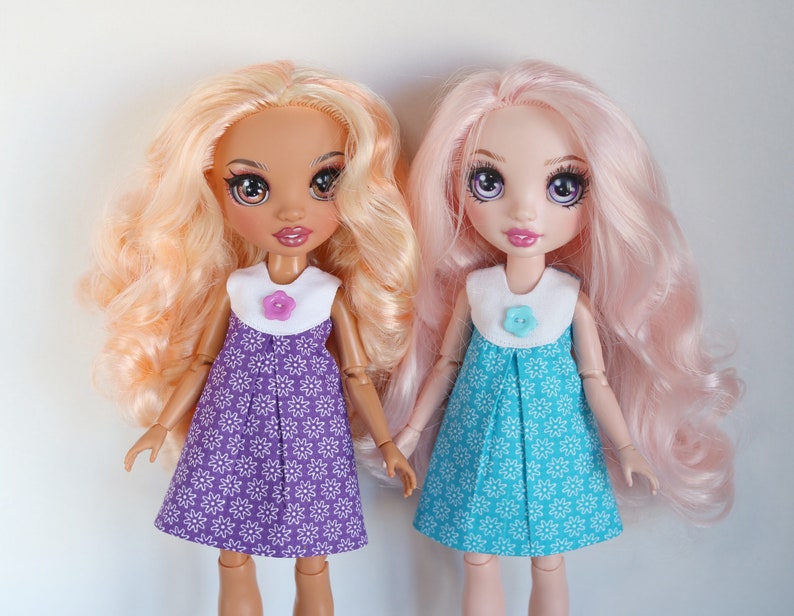May include: Two dolls wearing dresses with floral patterns. The doll on the left has long, wavy, light blonde hair and is wearing a purple dress with white flowers. The doll on the right has long, wavy, pink hair and is wearing a turquoise dress with white flowers.
