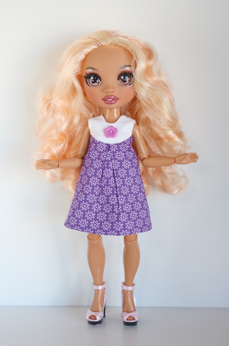 May include: A doll with long blonde hair wearing a purple dress with white flowers and a white collar with a purple flower button. The doll is wearing pink high heels.