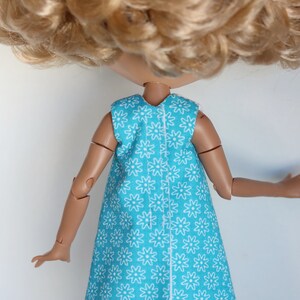 May include: A doll wearing a sleeveless dress with a white floral pattern on a turquoise background. The dress has a white seam down the back.