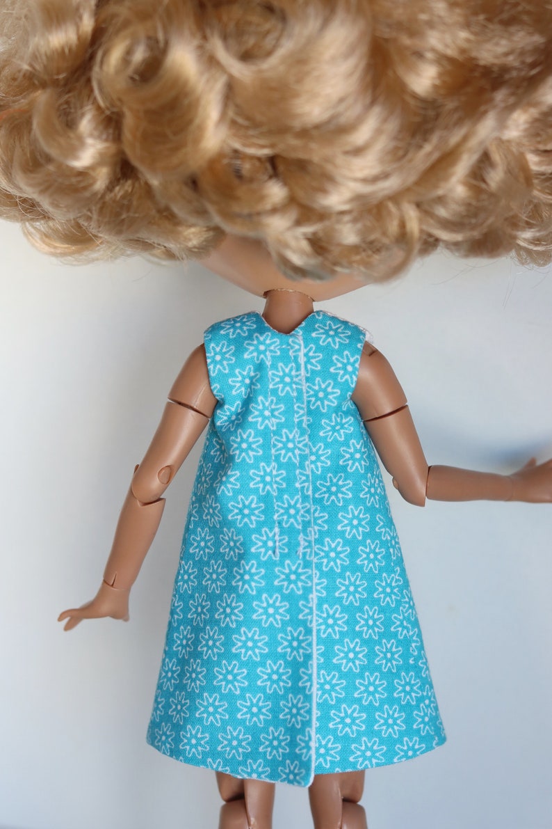 May include: A doll wearing a sleeveless dress with a white floral pattern on a turquoise background. The dress has a white seam down the back.