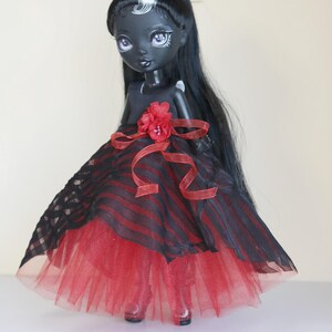 May include: A dark-skinned doll with long black hair and a red and black striped dress. The doll's dress has a red tulle skirt and a red flower accent at the waist. The doll has pale purple eyes and is wearing red shoes.