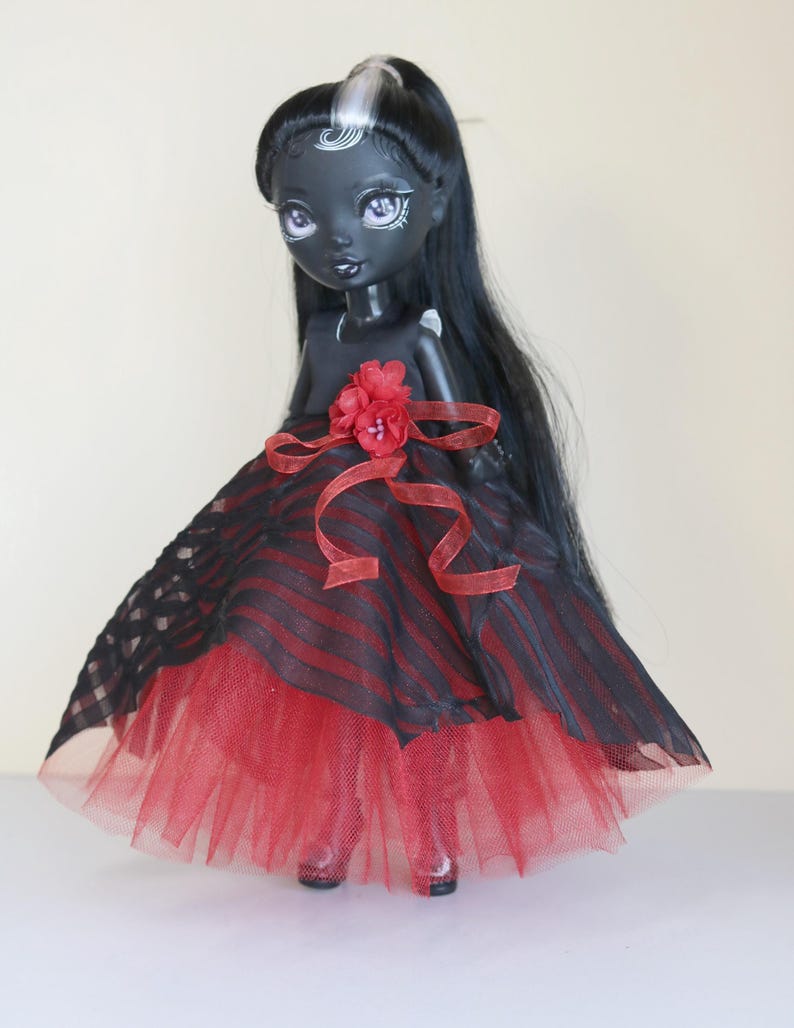 May include: A dark-skinned doll with long black hair and a red and black striped dress. The doll's dress has a red tulle skirt and a red flower accent at the waist. The doll has pale purple eyes and is wearing red shoes.