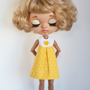 May include: A doll with blonde curly hair wearing a yellow dress with white floral print and a white collar with a yellow flower button.