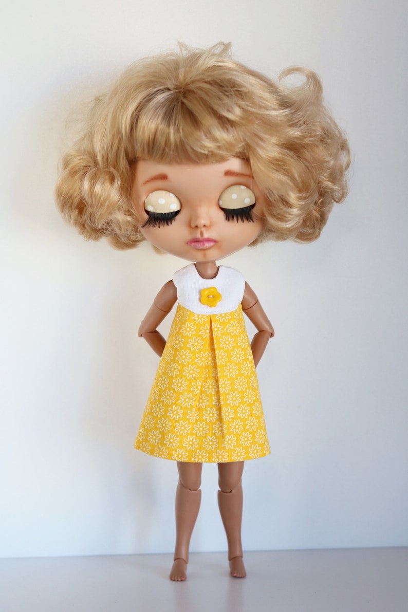 May include: A doll with blonde curly hair wearing a yellow dress with white floral print and a white collar with a yellow flower button.