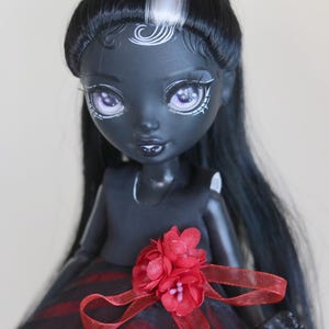May include: A doll with a dark complexion, black hair, and a red and black dress. The doll has white accents around the eyes and on the forehead. A red flower and ribbon adorn the dress.
