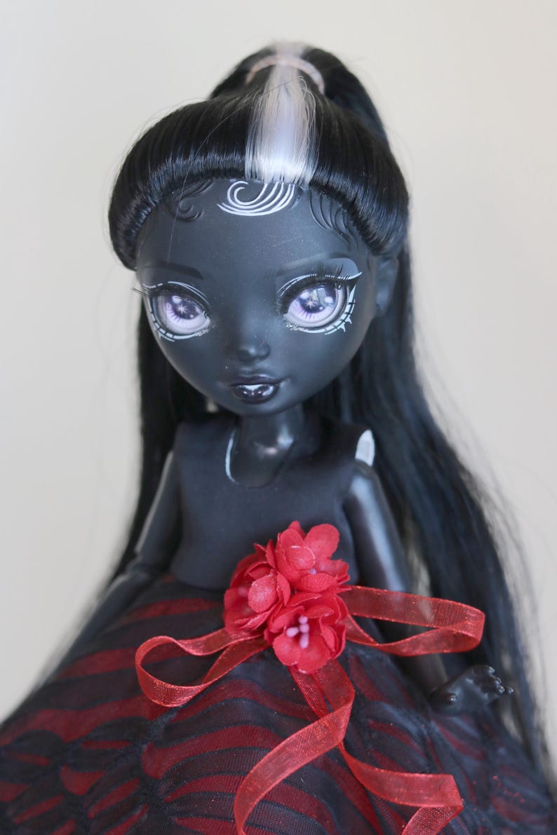 May include: A doll with a dark complexion, black hair, and a red and black dress. The doll has white accents around the eyes and on the forehead. A red flower and ribbon adorn the dress.